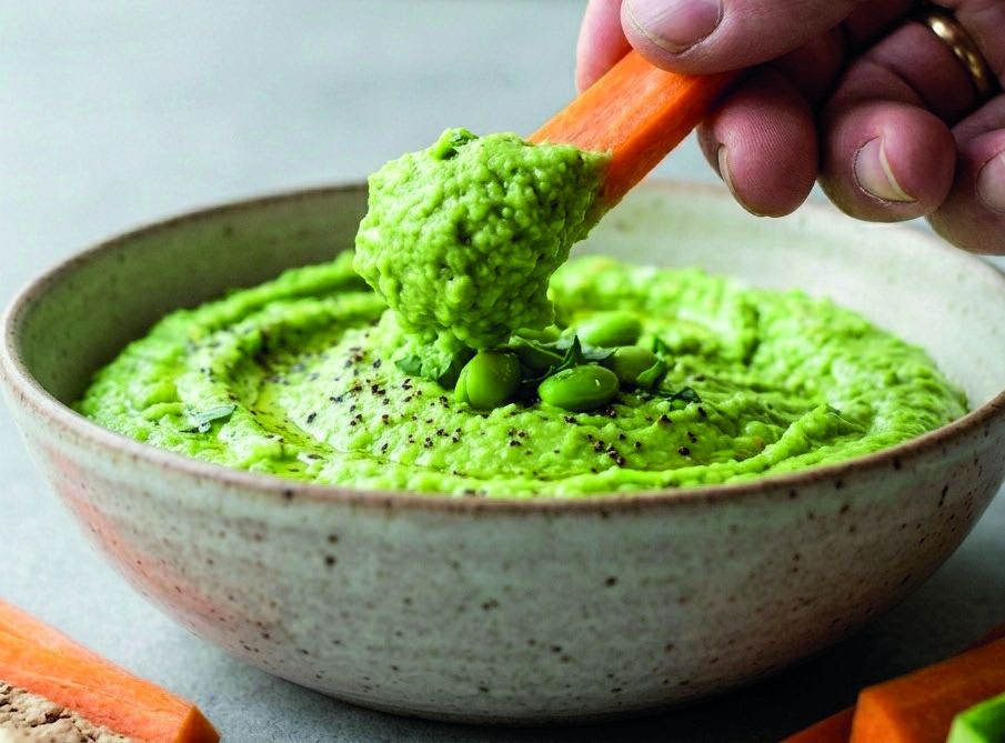 A carrot stick being dipped into a bowl of bright green edamame dip.