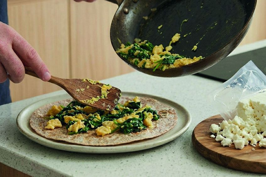 Scrambled egg and wilted spinach being spooned onto a wholemeal wrap with crumbled feta.